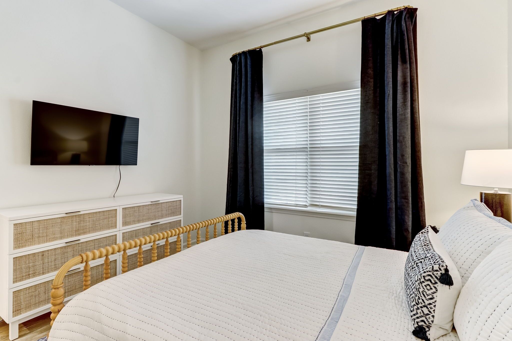 A bedroom with a bed, a TV mounted on the wall, a bedside table, and a window with curtains.