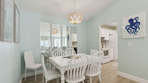 A dining area with a white table and chairs, a hanging light fixture, and a kitchen visible in the background. - North End Gem - Pet Friendly Fernandina Beach Cottage (Fernandina Beach)