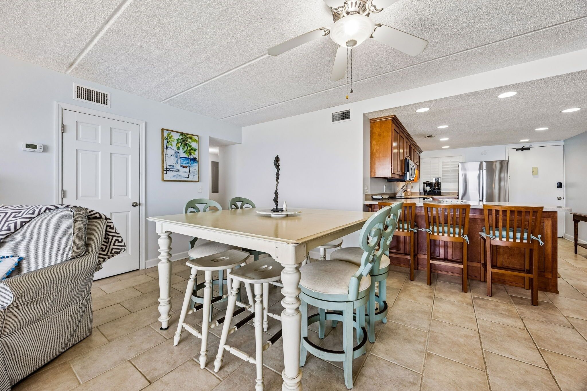 A dining area with a table and chairs, a kitchen in the background, and a ceiling fan.