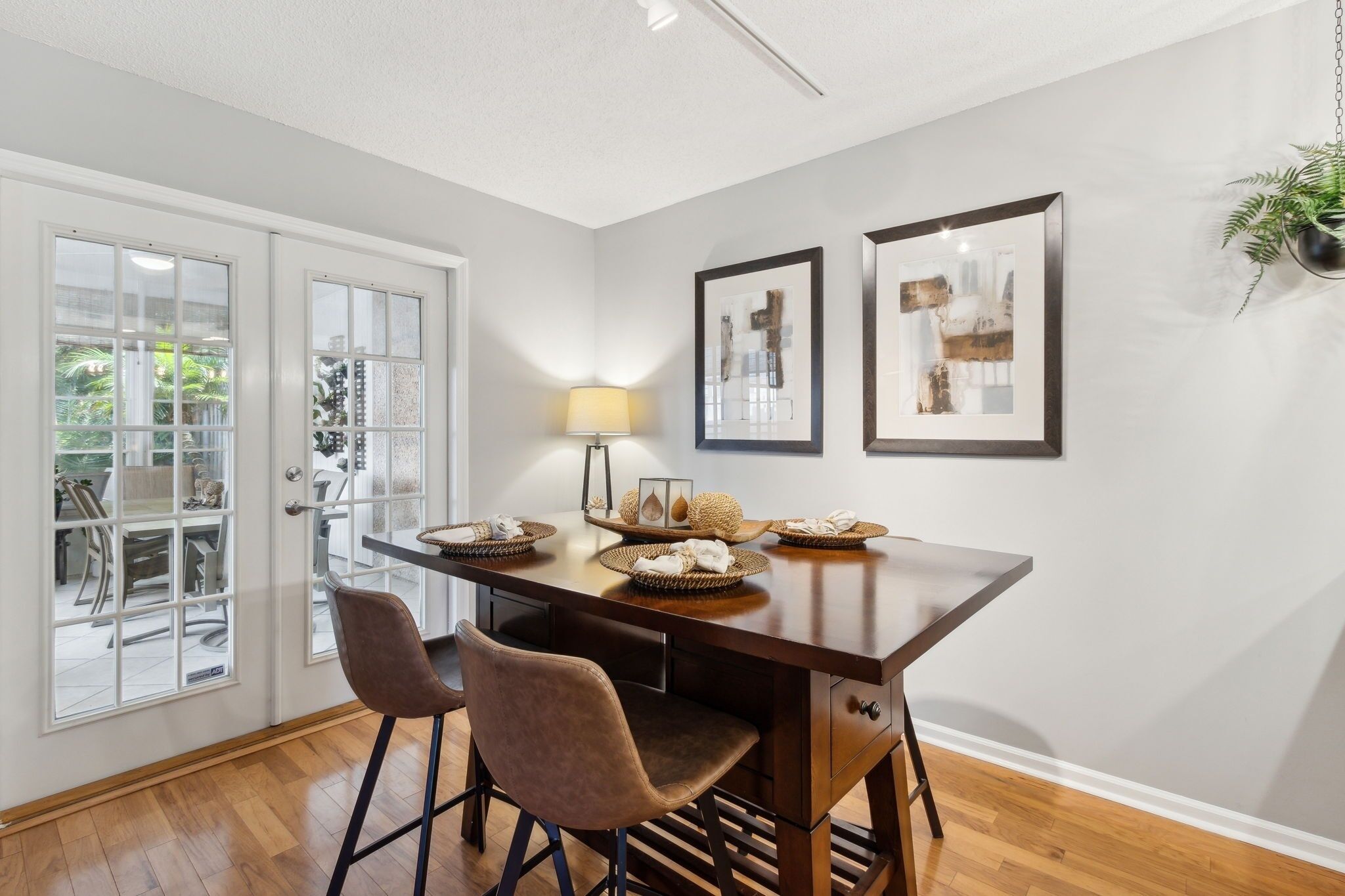 A dining area with a wooden table, two chairs, a lamp, and framed artwork on the wall.
