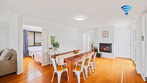 Amplia sala de estar con una mesa de comedor de madera, sillas blancas, una chimenea y vistas a un muro de ladrillo a través de una puerta abierta.