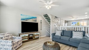 Een moderne woonkamer met een flatscreen televisie, een grijze bank, een gestreept fauteuil, een rond geweven pouf en een trap met een witte leuning.