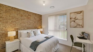 A hotel room with a brick wall, a bed with white linens, a desk with a laptop, and a window with blinds.