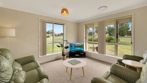 A spacious living room with a large TV, comfortable green leather sofas, a small round table, and large windows offering a view of the outdoors.