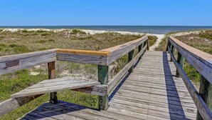 Un muelle de madera que conduce a una playa con dunas de arena y un cielo azul despejado.