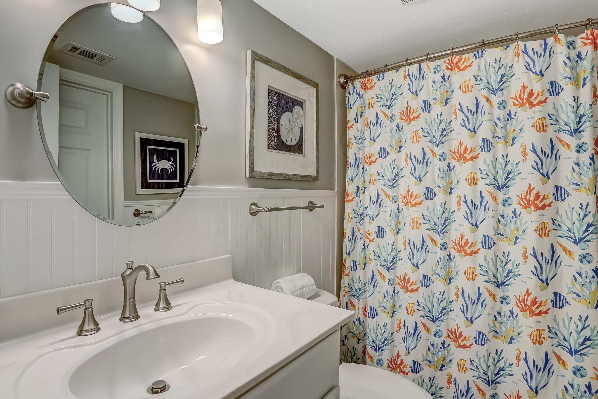 A bathroom with a white sink, three faucets, a mirror, a framed picture, and a shower curtain with a fish pattern.