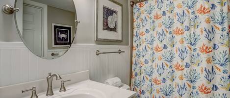 A bathroom with a white sink, three faucets, a mirror, a framed picture, and a shower curtain with a fish pattern.
