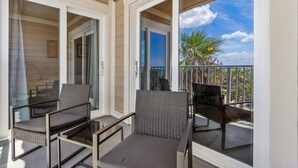 A balcony with two chairs and a table, glass doors, and a view of palm trees and a clear sky. - Vintage Ocean View Paradise - 1BR - Beachfront (Fernandina Beach)