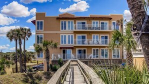 A multi-story hotel with balconies, a wooden walkway leading to the entrance, and palm trees in the background. - Vintage Ocean View Paradise - 1BR - Beachfront (Fernandina Beach)