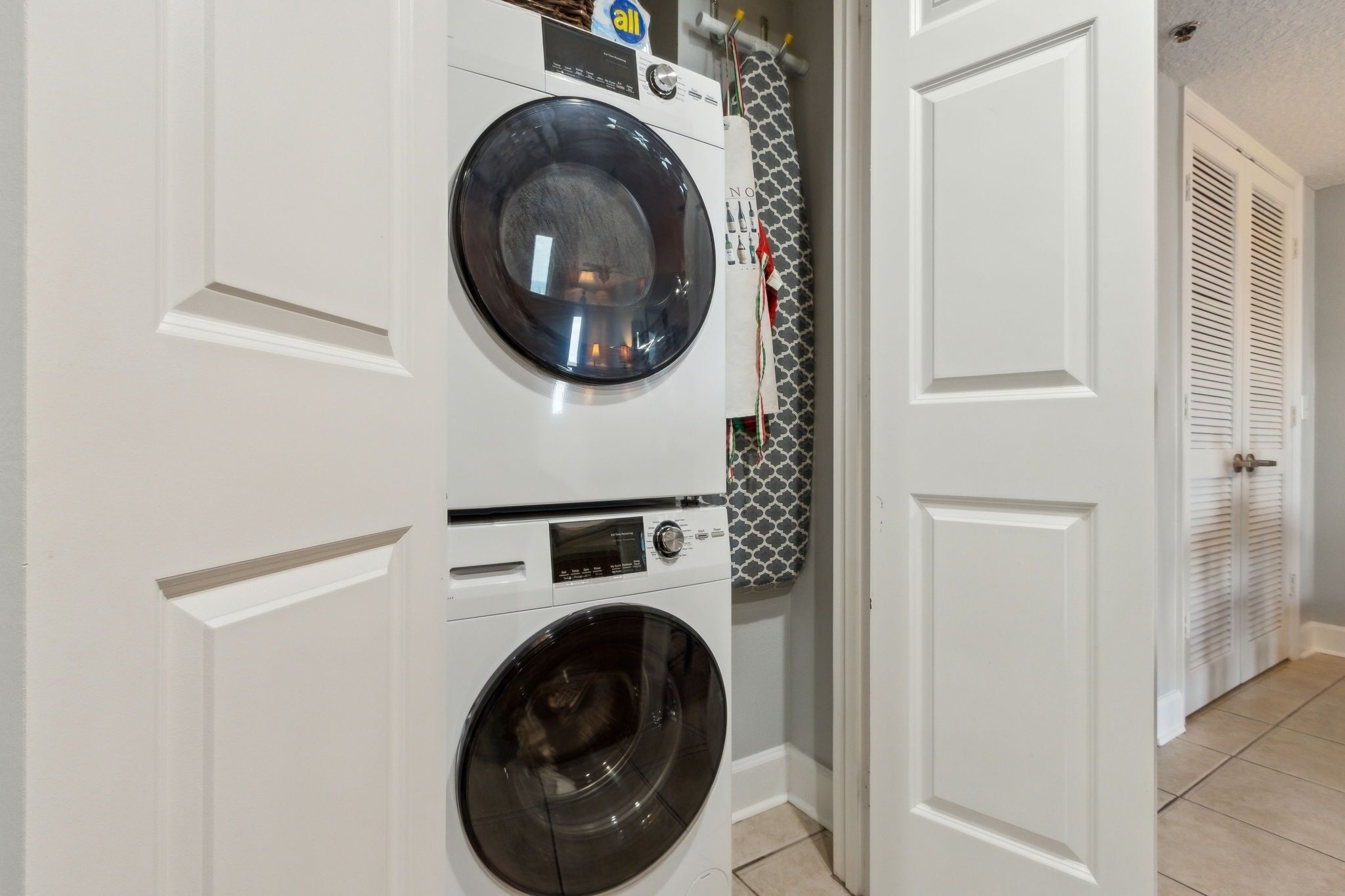 A laundry room with stacked washers and dryers, a patterned shower curtain, and a door with louvered shutters.