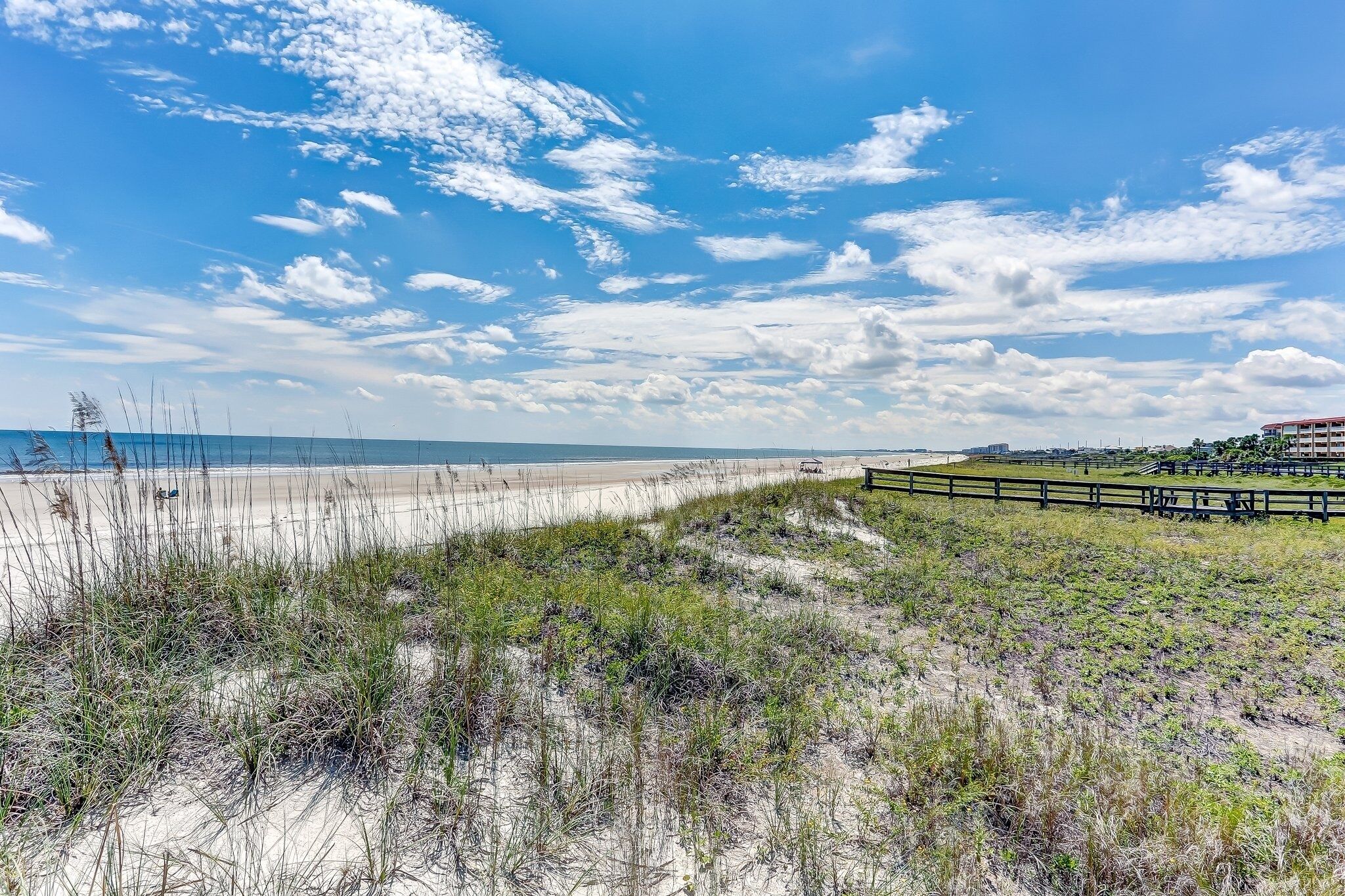 Une plage avec une clôture en bois, des dunes herbeuses et un ciel dégagé.