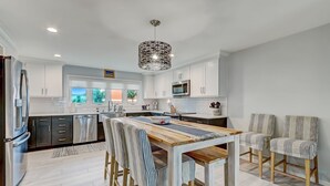 A modern kitchen with white cabinets, stainless steel appliances, and a wooden dining table with striped chairs. - Spacious Beachfront 3BR in Amelia Island Sandpiper Loop (Fernandina Beach)