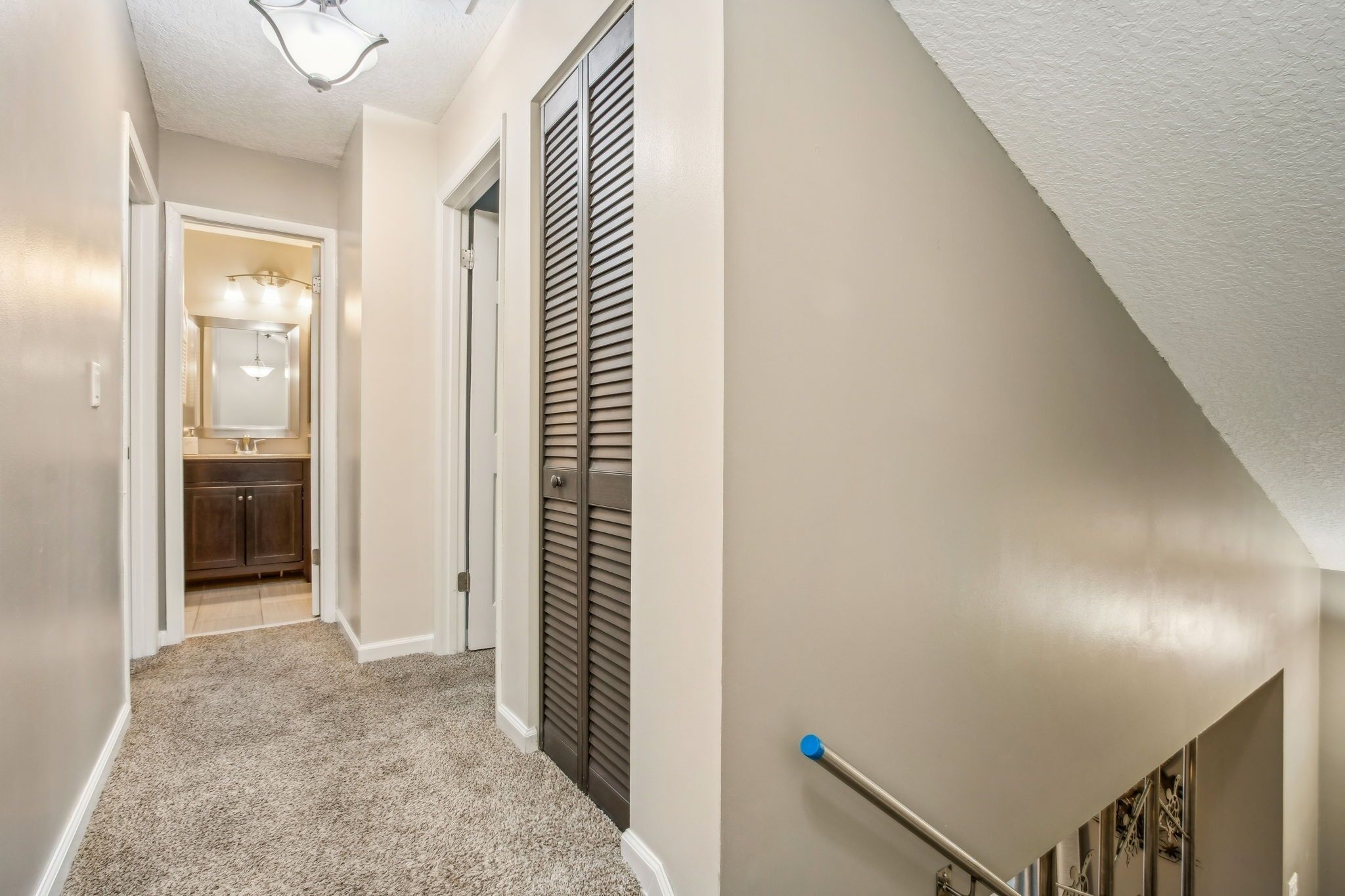 A hallway with a carpeted floor, a door leading to a bathroom, and a staircase with a handrail.