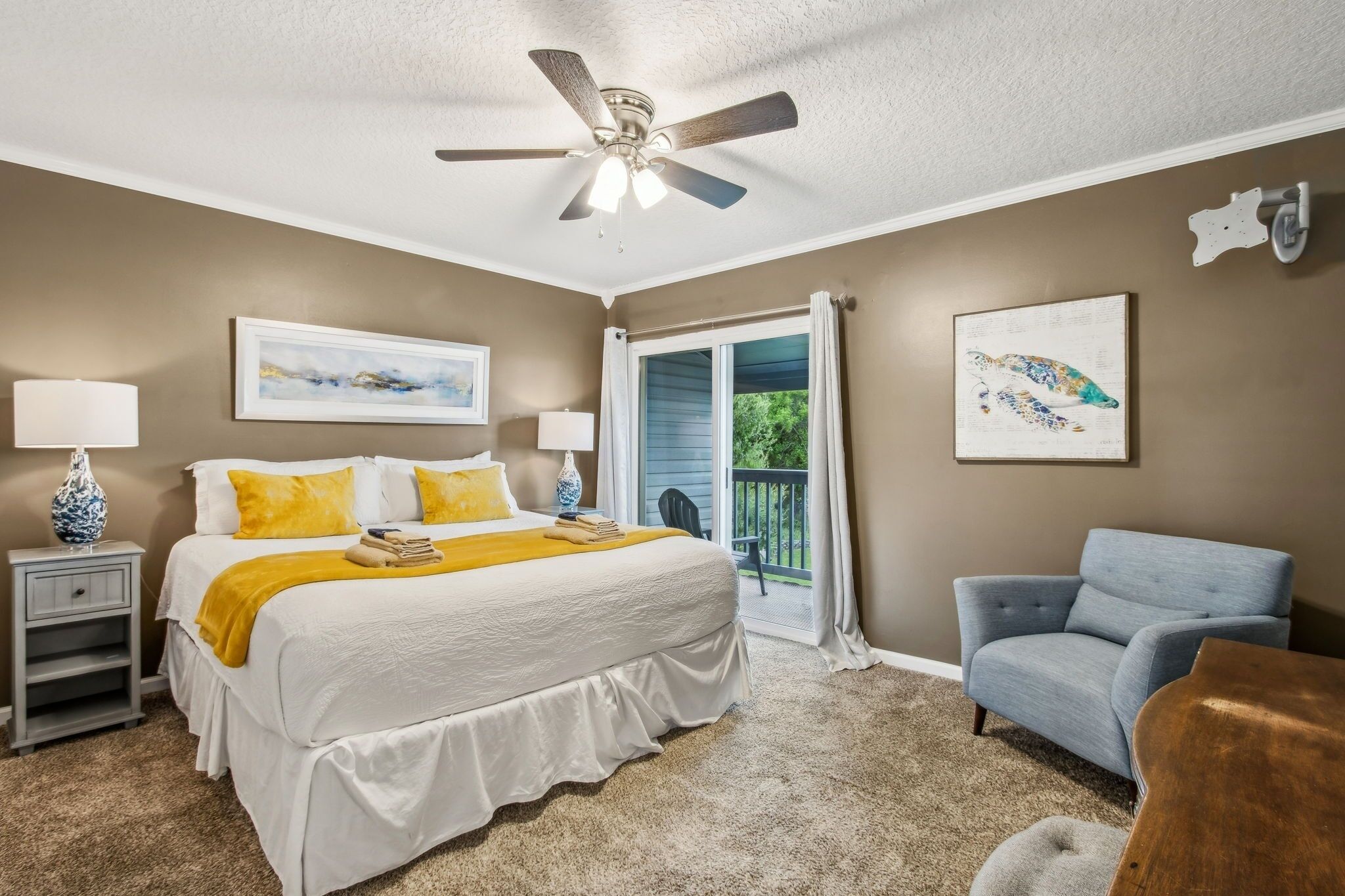 A bedroom with a bed, two bedside lamps, a ceiling fan, a chair, and a balcony with a view.