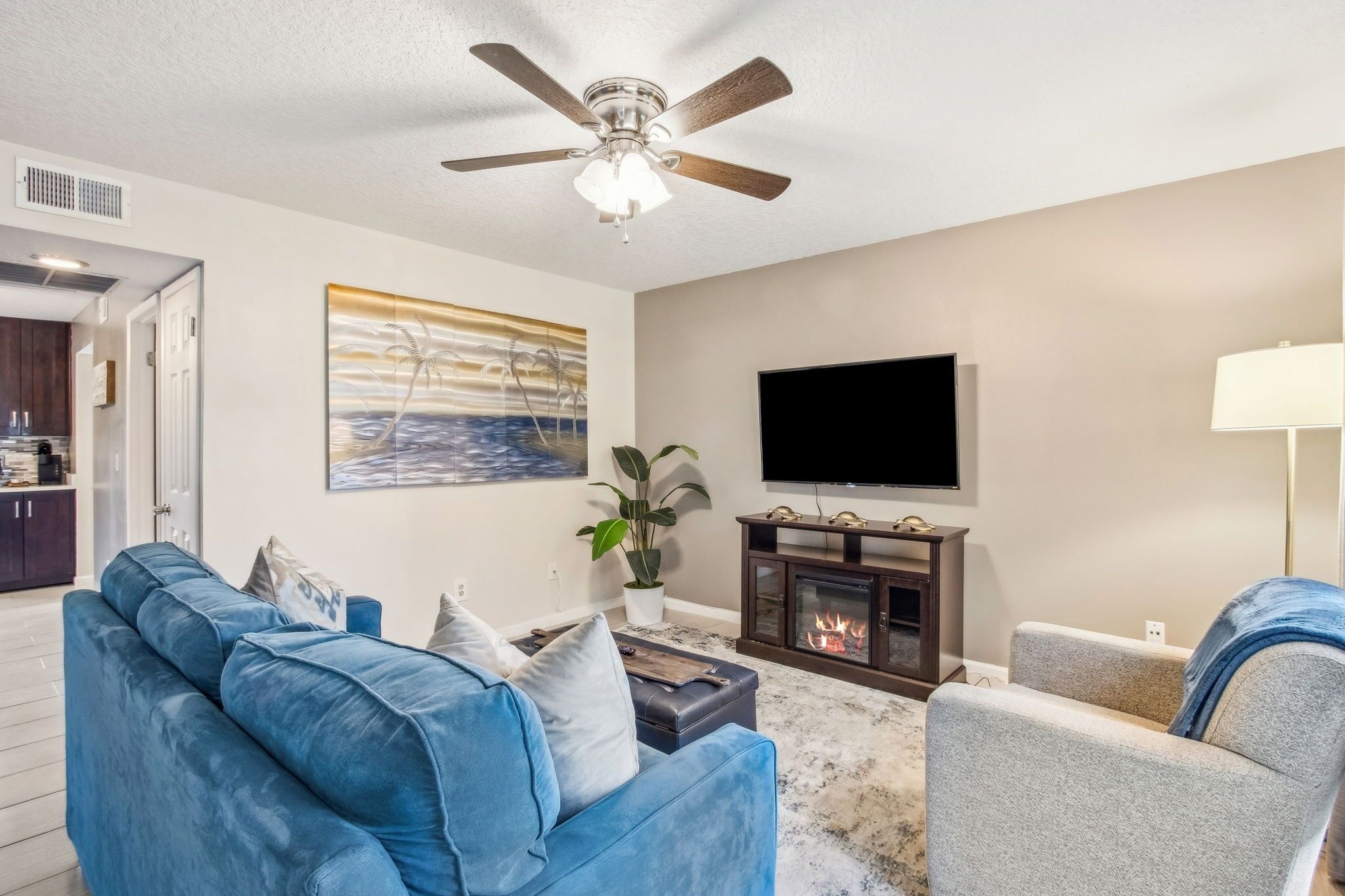 A living room with a blue sofa, a grey armchair, a flat-screen TV, a fireplace, and a ceiling fan.