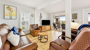 A living room with a brown leather sofa, a wooden coffee table, and a television on a wooden cabinet. There is a painting on the wall and a window with blinds. - Spacious Beachfront Duplex - Top Floor - Dog Friendly 124 S Fletcher (Fernandina Beach)