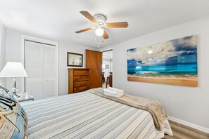 Une chambre à coucher avec un lit, un ventilateur de plafond, un tableau représentant une plage et une commode en bois.