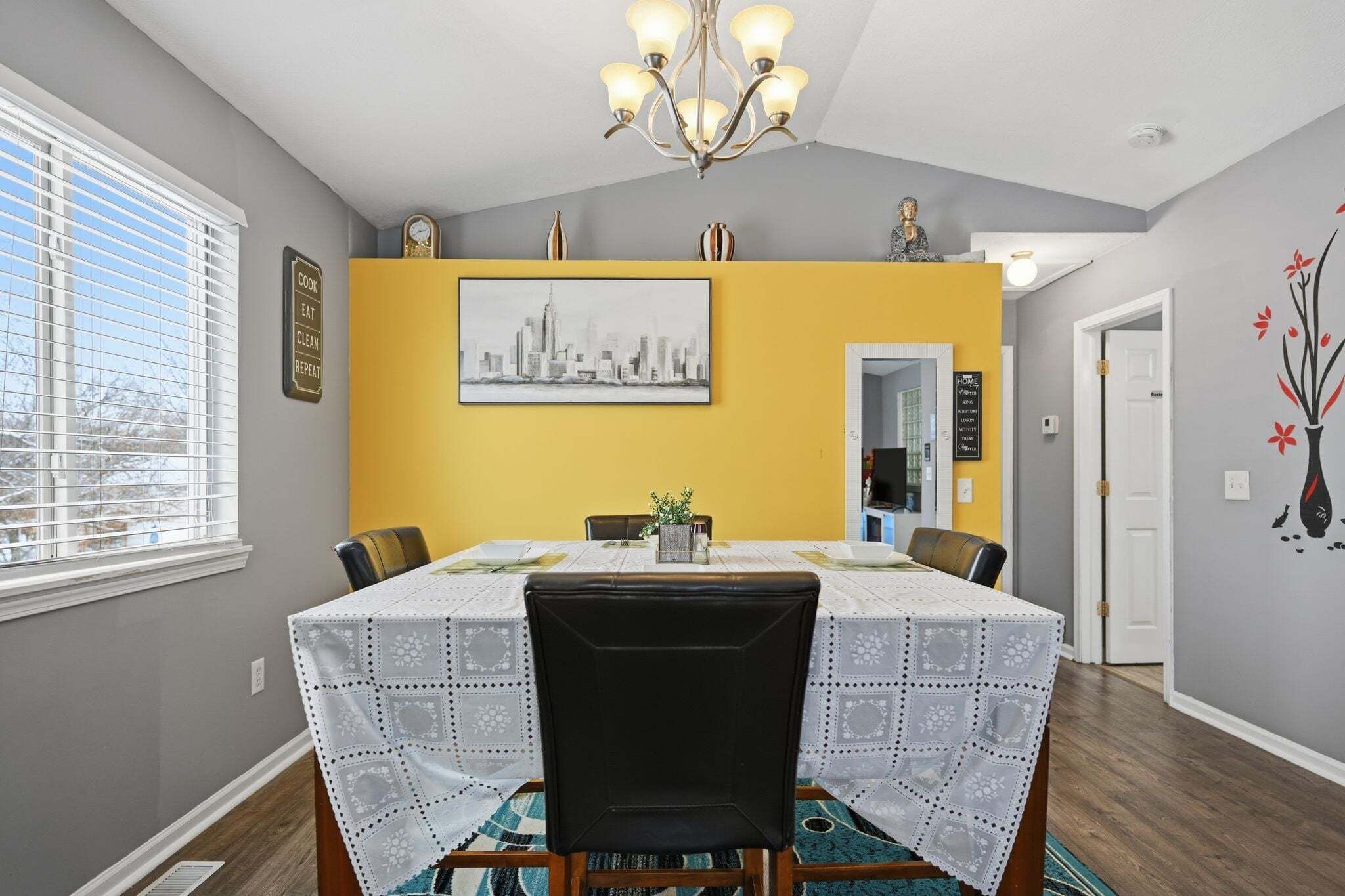A dining area with a table set for four, a chandelier, a cityscape painting, and a door leading to another room.