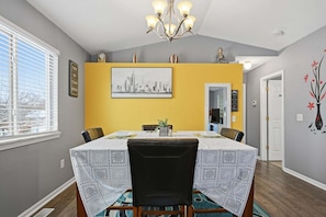 A dining area with a table set for four, a chandelier, a cityscape painting, and a door leading to another room. - Pet Friendly Lansing Home - Private Gym - Near MSU (Lansing)