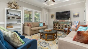 A living room with a beige sofa, blue armchair, and a coffee table. There is a television mounted on the wall, a ceiling fan, and a bookshelf with decorative items. - Bienville Bungalow - 304 Bienville Blvd 2 Bedroom Home (Dauphin Island)