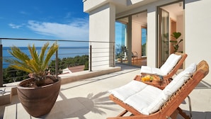 A balcony with two wooden lounge chairs, a potted plant, and a table with a fruit bowl and drinks.