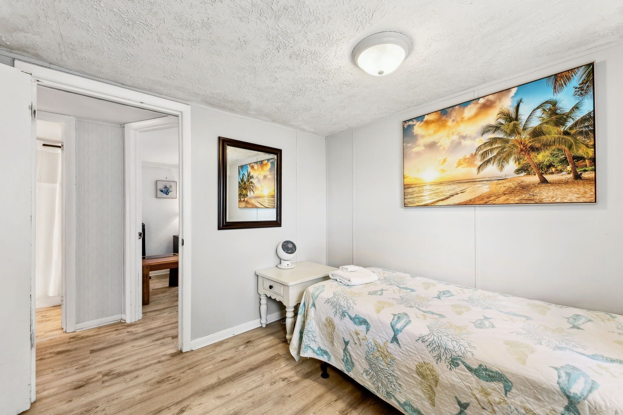 Une chambre à coucher avec un lit, une table de chevet avec une lampe, un cadre photo et un tableau représentant une scène de plage.