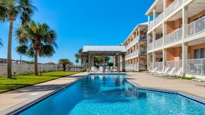 A swimming pool with a covered area, palm trees, and a building with balconies. - Dauphin Luv B - Beach Club #201-b 2 Bedroom Condo (Dauphin Island)