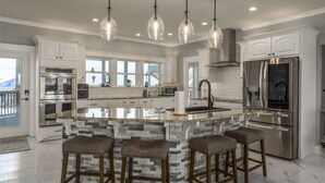 A modern kitchen with white cabinets, a large island with stools, stainless steel appliances, and pendant lighting. - Bay Breeze - 2420 Bienville 4 Bedroom Home (Dauphin Island)