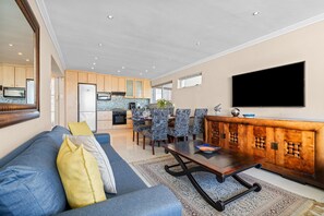 A living room with a sofa, a coffee table, a dining area with chairs, a kitchen with cabinets, and a flat-screen TV mounted on the wall. - Sailaway Sea Point (Cape Town)