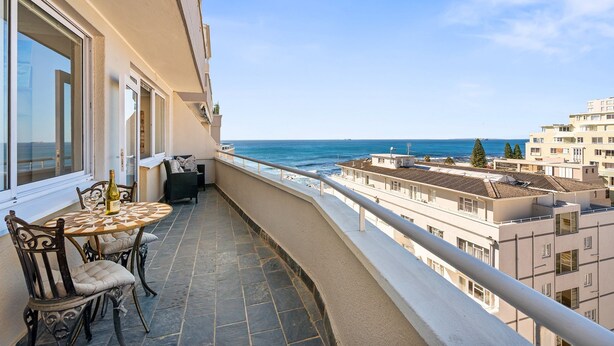 Un balcón con una mesa y sillas, con vistas a la costa y edificios al fondo.