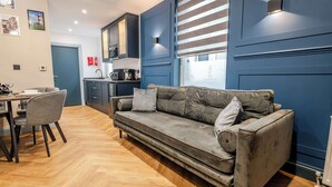 Ein modernes Wohnzimmer mit einem grauen Sofa, einem Esstisch mit Stühlen, einer Küchenzeile und einem großen Fenster mit Jalousien.