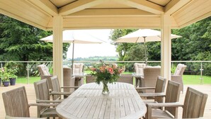 A covered patio with a wooden table and wicker chairs, surrounded by potted plants and umbrellas. - The Hayloft (Falmouth)