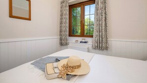 A bedroom with a bed, a window with curtains, a radiator, a mirror, and a small table with a book and a teapot. - The Hayloft (Falmouth)