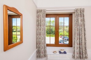 A room with a wooden-framed window, patterned curtains, a small shelf with a teapot and book, and a mirror on the wall. - The Hayloft (Falmouth)