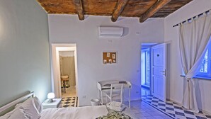 Une chambre à coucher comprenant un lit, une table de chevet avec une lampe, un petit bureau, une chaise, un climatiseur fixé au mur et une porte donnant sur une autre pièce.