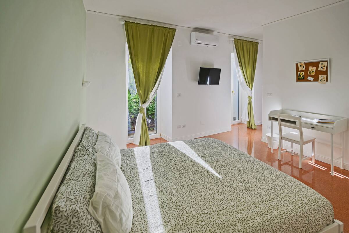 Ein Schlafzimmer mit Bett, Schreibtisch und Stuhl. Ein Fenster mit Vorhängen, ein wandmontierter Fernseher und ein Korkbrett mit Bildern sind ebenfalls vorhanden.