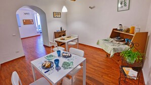 Un coin repas avec une table blanche dressée pour deux personnes, un plancher en bois et, au fond, une petite cuisine.