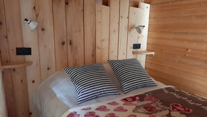 A wooden room with a bed, two pillows, a white blanket, and a red patterned throw.