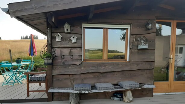 A rustic cabin with a wooden exterior, a window with a view, a bench with cushions, and a patio area with outdoor furniture.