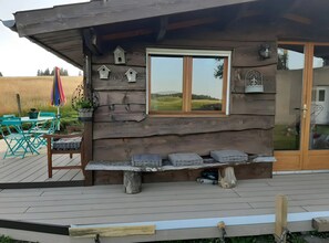 A rustic cabin with a wooden exterior, a window with a view, a bench with cushions, and a patio area with outdoor furniture.