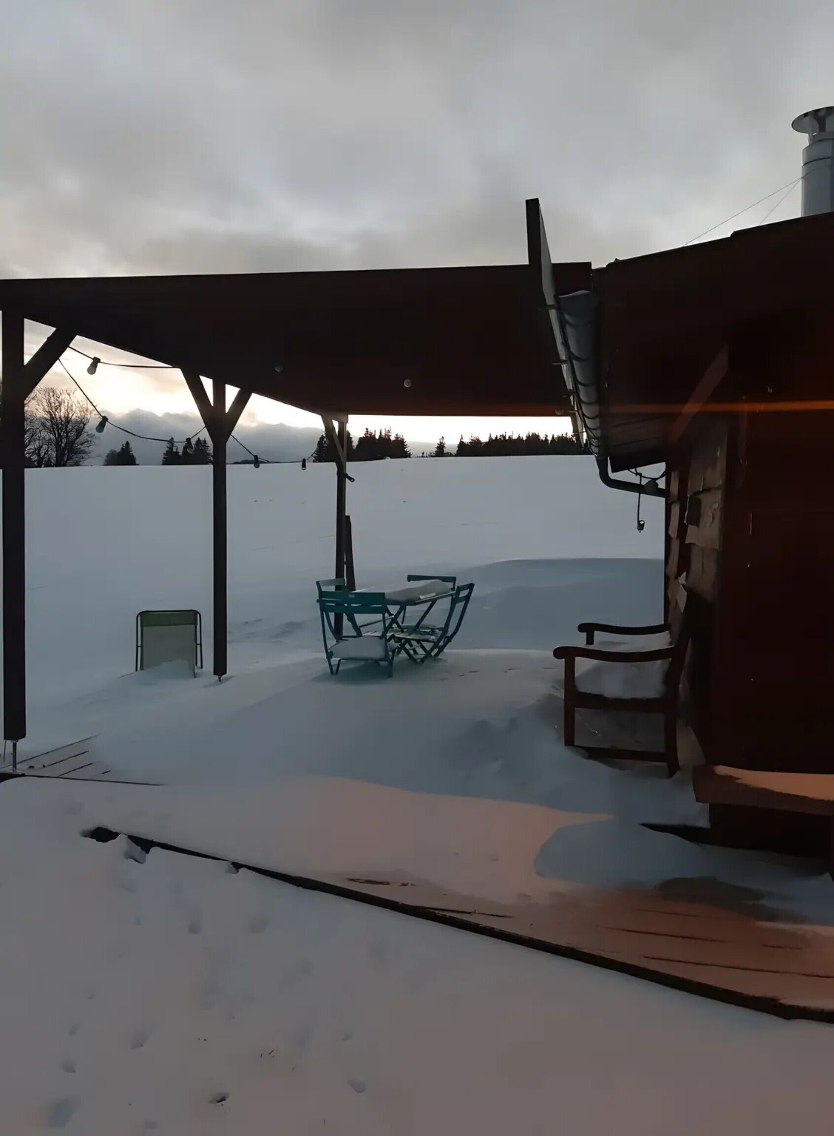 Et overdækket udendørs område med et bord og stole, en mindre bygning og et sneklædt landskab.