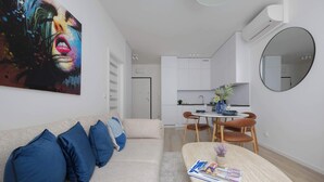 A modern living room with a beige sofa, blue cushions, a coffee table with magazines, a large abstract painting, a dining area with a round table and chairs, and a kitchen with white cabinets. - Stylish Apartment with Balcony | Air Conditioning | Close to the Metro (Warszawa)