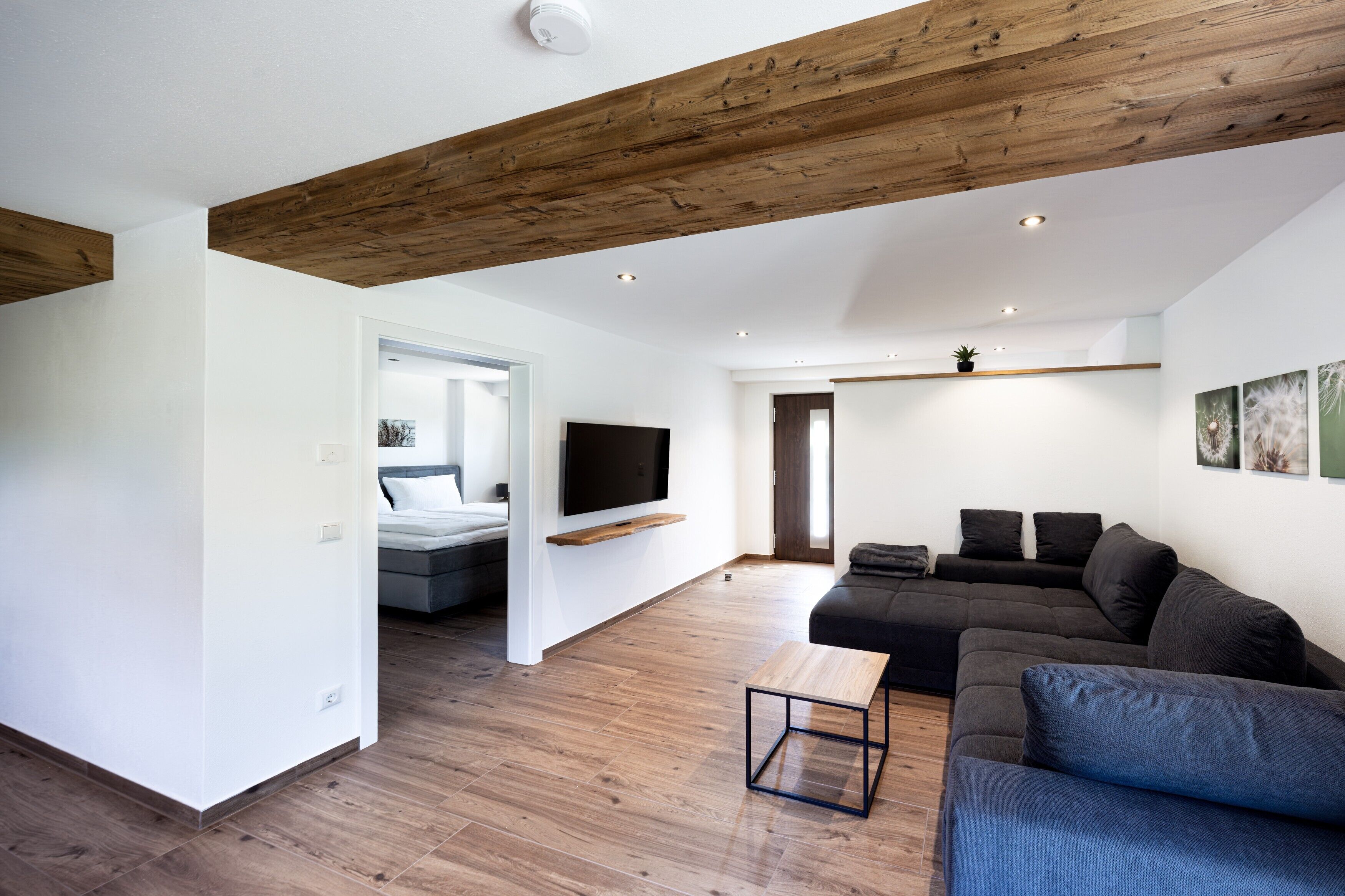 A modern living room with a wooden ceiling, a grey sofa, a wooden coffee table, and a flat-screen TV mounted on the wall.