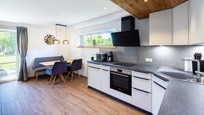 A modern kitchen with a Bosch oven, a dining area with a bench and chairs, and a view of greenery outside. - Holiday Apartment for 2 with Private Terrace, Shared Garden & Wi-Fi at Ferienhaus Waldherr (Waging am See)