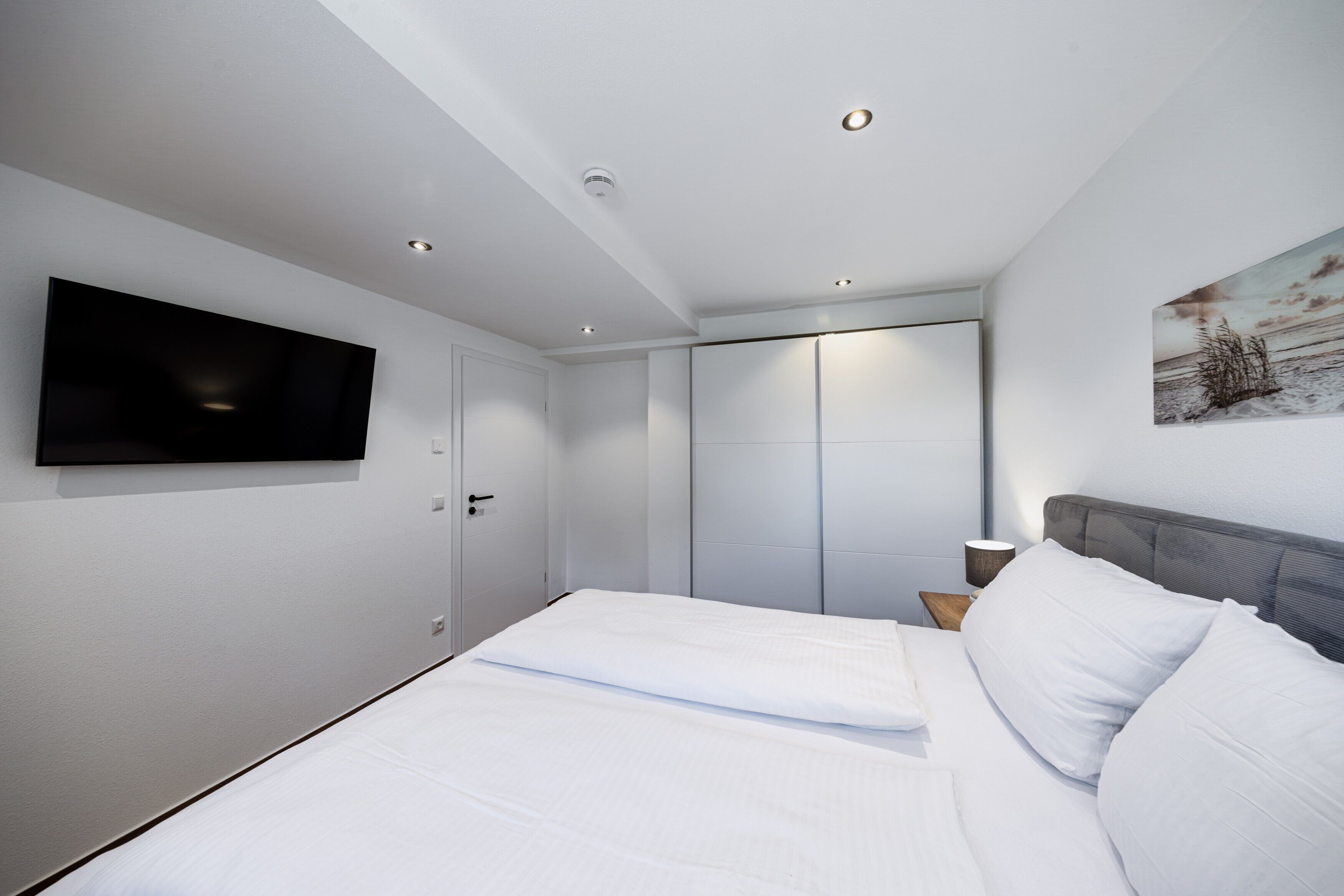 Ein modernes Schlafzimmer mit Wand-Fernseher, einem Bett mit weißen Bettwäsche, einem Nachttisch mit Lampe und einem Kleiderschrank mit Schiebetüren.
