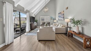 A spacious living room with a large window, a sofa, a coffee table, and a TV. - Beach Condo~Free Golf Cart~3BD~2BA~Sleeps 8~Balcony~Walk to Beach~Pool (Miramar Beach)