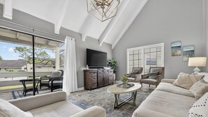 A spacious living room with a large TV, comfortable seating, a balcony with outdoor furniture, and a view of the neighborhood. - Beach Condo~Free Golf Cart~3BD~2BA~Sleeps 8~Balcony~Walk to Beach~Pool (Miramar Beach)