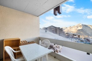Un balcón con una mesa y sillas, un estante de madera, y vistas a una montaña nevada y edificios.