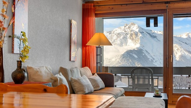 Habitación de hotel con sofá, una mesa, lámpara, un jarrón con flores y vistas a montañas nevadas.