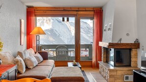 Una sala de estar acogedora con chimenea, un sofá, una mesa y vistas a montañas a través de amplios ventanales.